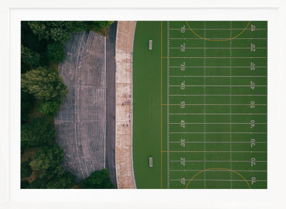 An aerial top-down photograph of an American football field beside empty, weathered concrete stadium bleachers. The vibrant green field is marked with white yard lines, and the gray stands are nestled amongst lush green trees. The entire scene is enclosed in a silver frame. Artwork