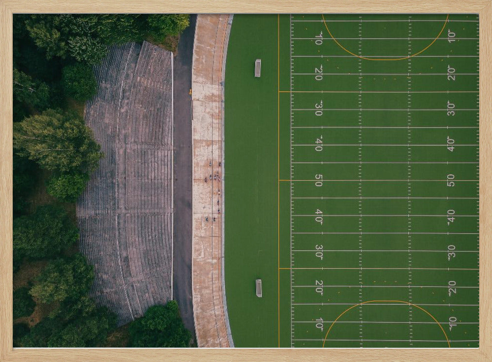 An aerial top-down photograph of an American football field beside empty, weathered concrete stadium bleachers. The vibrant green field is marked with white yard lines, and the gray stands are nestled amongst lush green trees. The entire scene is enclosed in a silver frame. Artwork