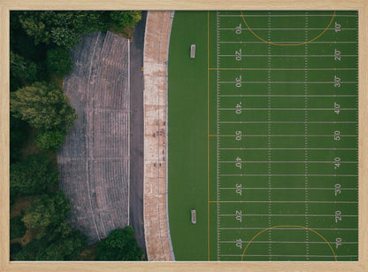 An aerial top-down photograph of an American football field beside empty, weathered concrete stadium bleachers. The vibrant green field is marked with white yard lines, and the gray stands are nestled amongst lush green trees. The entire scene is enclosed in a silver frame. Artwork