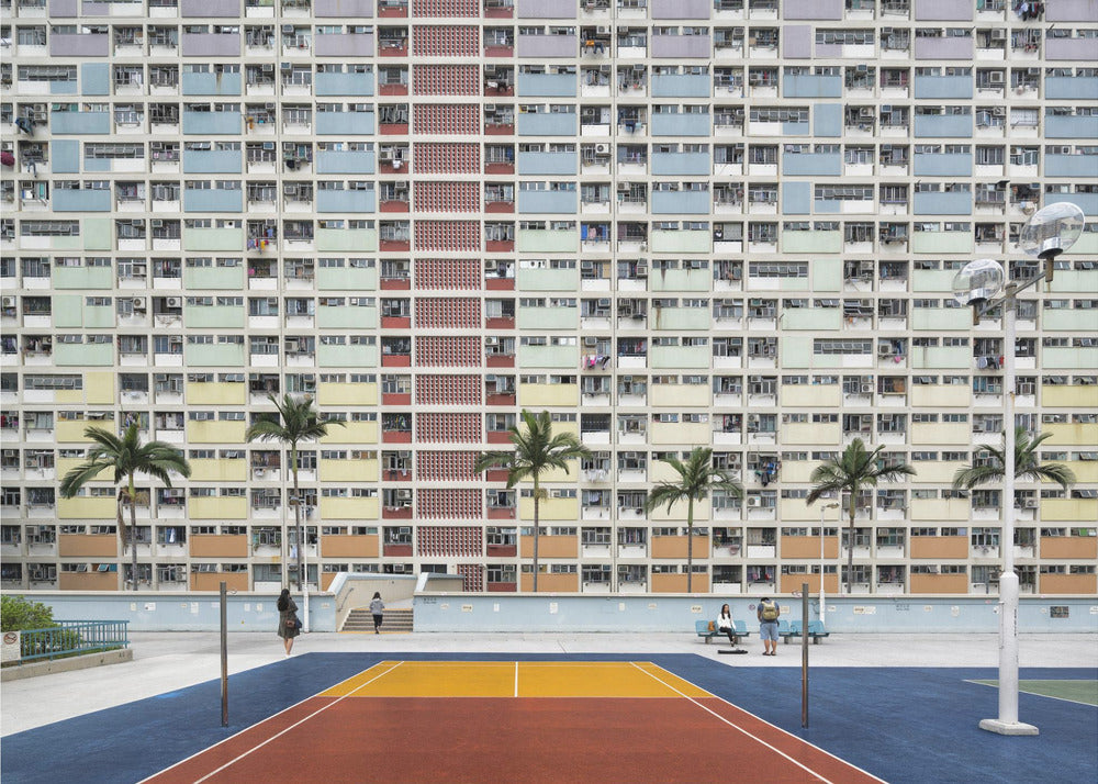 A wide, symmetrical photograph of the colorful Choi Hung Estate apartment building in Hong Kong, featuring a facade with a repeating pattern of pastel blue, green, yellow, and orange blocks. In front of the building is a vibrant basketball court painted in bold sections of blue, red, and yellow, with several palm trees lining the courtyard. Print