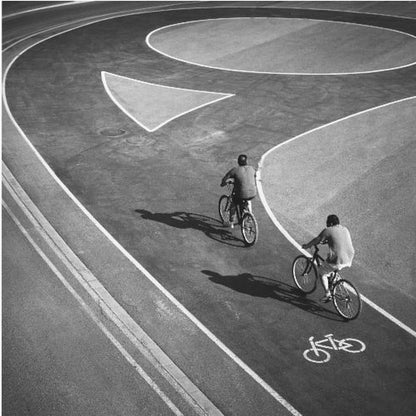 A framed, high-angle black and white photograph of two people cycling on a curving asphalt bike path. The path is marked with bold white lines, including a large arrow and a bicycle symbol, creating a strong graphic pattern. The cyclists cast long shadows on the pavement. Poster