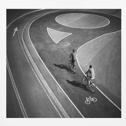 A framed, high-angle black and white photograph of two people cycling on a curving asphalt bike path. The path is marked with bold white lines, including a large arrow and a bicycle symbol, creating a strong graphic pattern. The cyclists cast long shadows on the pavement. Poster