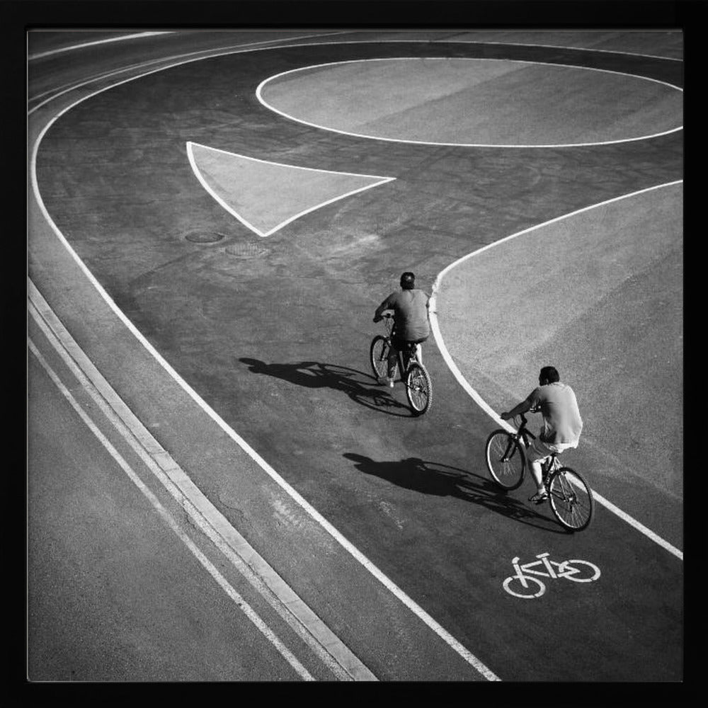 A framed, high-angle black and white photograph of two people cycling on a curving asphalt bike path. The path is marked with bold white lines, including a large arrow and a bicycle symbol, creating a strong graphic pattern. The cyclists cast long shadows on the pavement. Poster