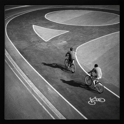 A framed, high-angle black and white photograph of two people cycling on a curving asphalt bike path. The path is marked with bold white lines, including a large arrow and a bicycle symbol, creating a strong graphic pattern. The cyclists cast long shadows on the pavement. Poster