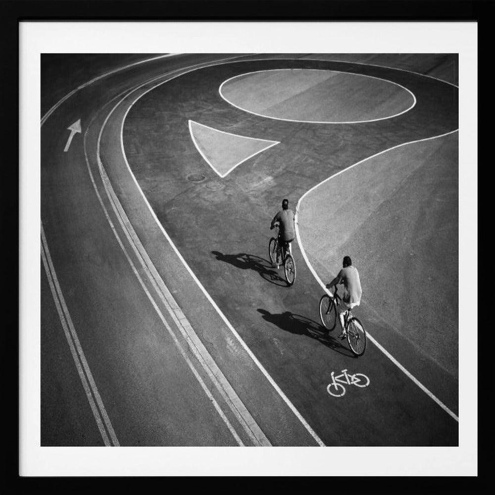 A framed, high-angle black and white photograph of two people cycling on a curving asphalt bike path. The path is marked with bold white lines, including a large arrow and a bicycle symbol, creating a strong graphic pattern. The cyclists cast long shadows on the pavement. Poster