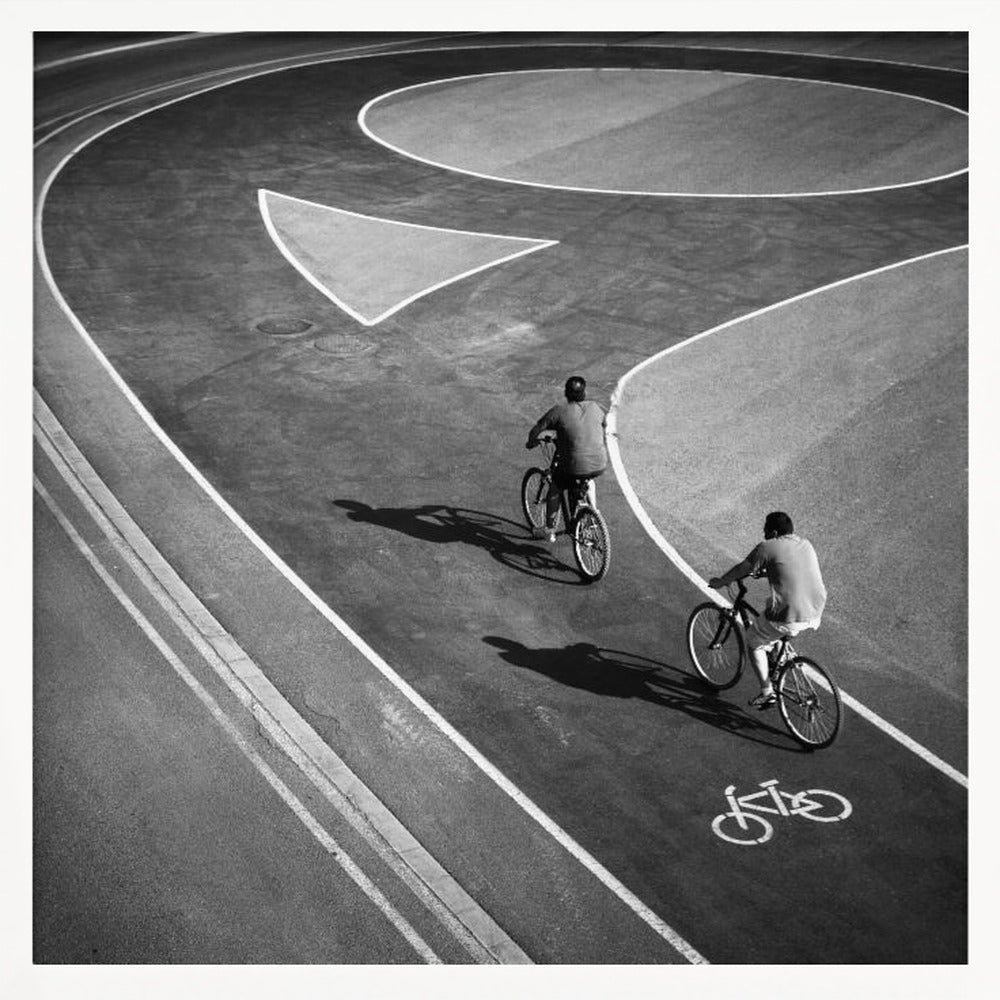 A framed, high-angle black and white photograph of two people cycling on a curving asphalt bike path. The path is marked with bold white lines, including a large arrow and a bicycle symbol, creating a strong graphic pattern. The cyclists cast long shadows on the pavement. Poster