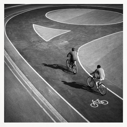 A framed, high-angle black and white photograph of two people cycling on a curving asphalt bike path. The path is marked with bold white lines, including a large arrow and a bicycle symbol, creating a strong graphic pattern. The cyclists cast long shadows on the pavement. Poster