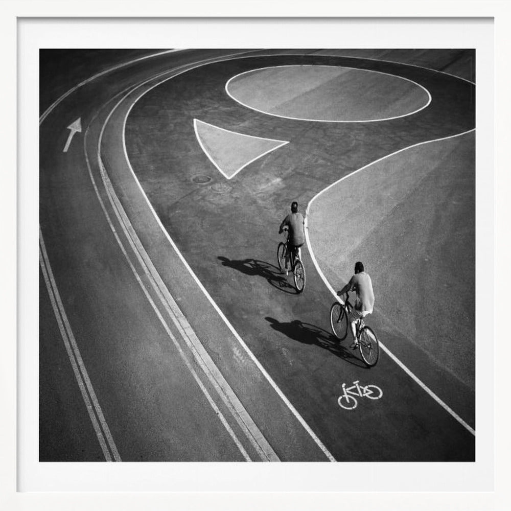 A framed, high-angle black and white photograph of two people cycling on a curving asphalt bike path. The path is marked with bold white lines, including a large arrow and a bicycle symbol, creating a strong graphic pattern. The cyclists cast long shadows on the pavement. Poster