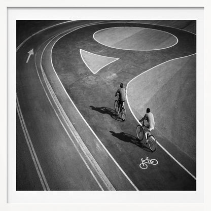 A framed, high-angle black and white photograph of two people cycling on a curving asphalt bike path. The path is marked with bold white lines, including a large arrow and a bicycle symbol, creating a strong graphic pattern. The cyclists cast long shadows on the pavement. Poster