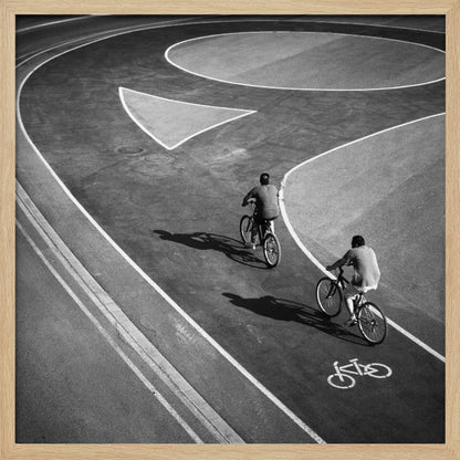 A framed, high-angle black and white photograph of two people cycling on a curving asphalt bike path. The path is marked with bold white lines, including a large arrow and a bicycle symbol, creating a strong graphic pattern. The cyclists cast long shadows on the pavement. Poster