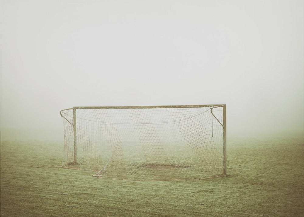 A framed photograph of an empty soccer goal with a net, standing alone on a green grass field enveloped in thick, white fog, creating a minimalist and atmospheric scene. Poster