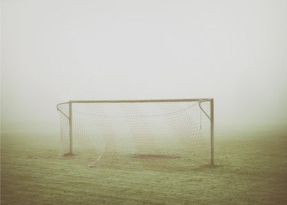 A framed photograph of an empty soccer goal with a net, standing alone on a green grass field enveloped in thick, white fog, creating a minimalist and atmospheric scene. Poster
