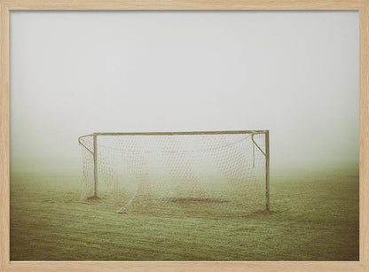 A framed photograph of an empty soccer goal with a net, standing alone on a green grass field enveloped in thick, white fog, creating a minimalist and atmospheric scene. Poster