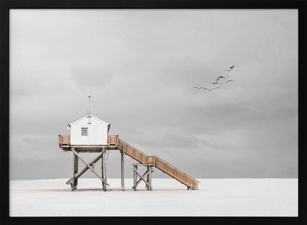 A minimalist, desaturated photograph of a small white cabin on tall wooden stilts on a vast, empty beach. A long wooden staircase leads up to the cabin. The sky is a flat, overcast grey, and a flock of dark birds is visible flying in the distance. Print