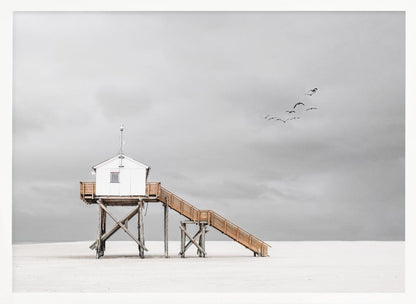 A minimalist, desaturated photograph of a small white cabin on tall wooden stilts on a vast, empty beach. A long wooden staircase leads up to the cabin. The sky is a flat, overcast grey, and a flock of dark birds is visible flying in the distance. Print