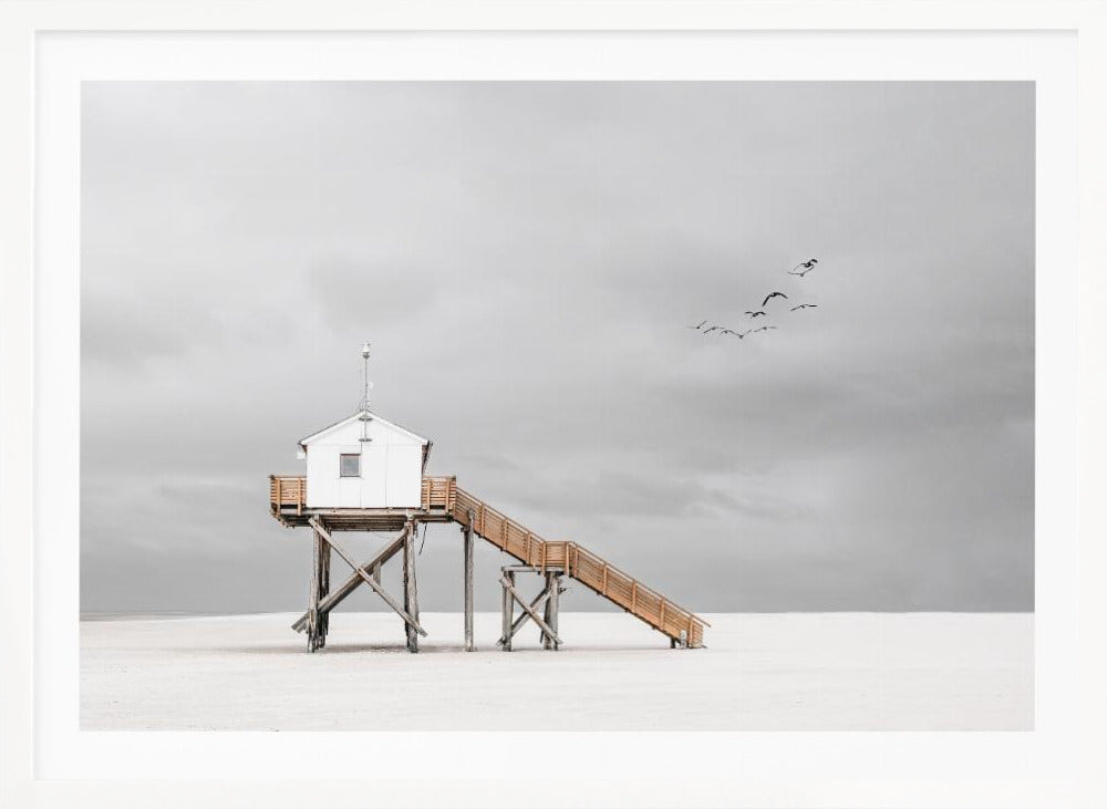 A minimalist, desaturated photograph of a small white cabin on tall wooden stilts on a vast, empty beach. A long wooden staircase leads up to the cabin. The sky is a flat, overcast grey, and a flock of dark birds is visible flying in the distance. Print