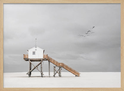 A minimalist, desaturated photograph of a small white cabin on tall wooden stilts on a vast, empty beach. A long wooden staircase leads up to the cabin. The sky is a flat, overcast grey, and a flock of dark birds is visible flying in the distance. Print