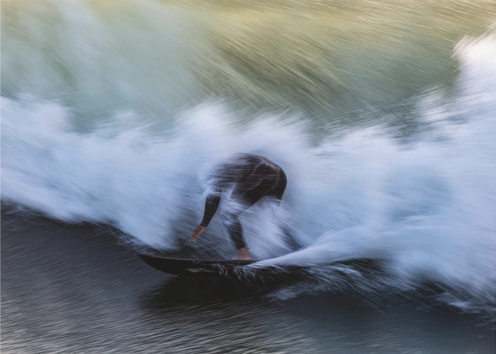 An abstract, motion-blur photograph of a surfer in a black wetsuit, crouched on their board and surrounded by a dynamic swirl of white sea spray and dark blue water, all enclosed in a silver frame. Artwork