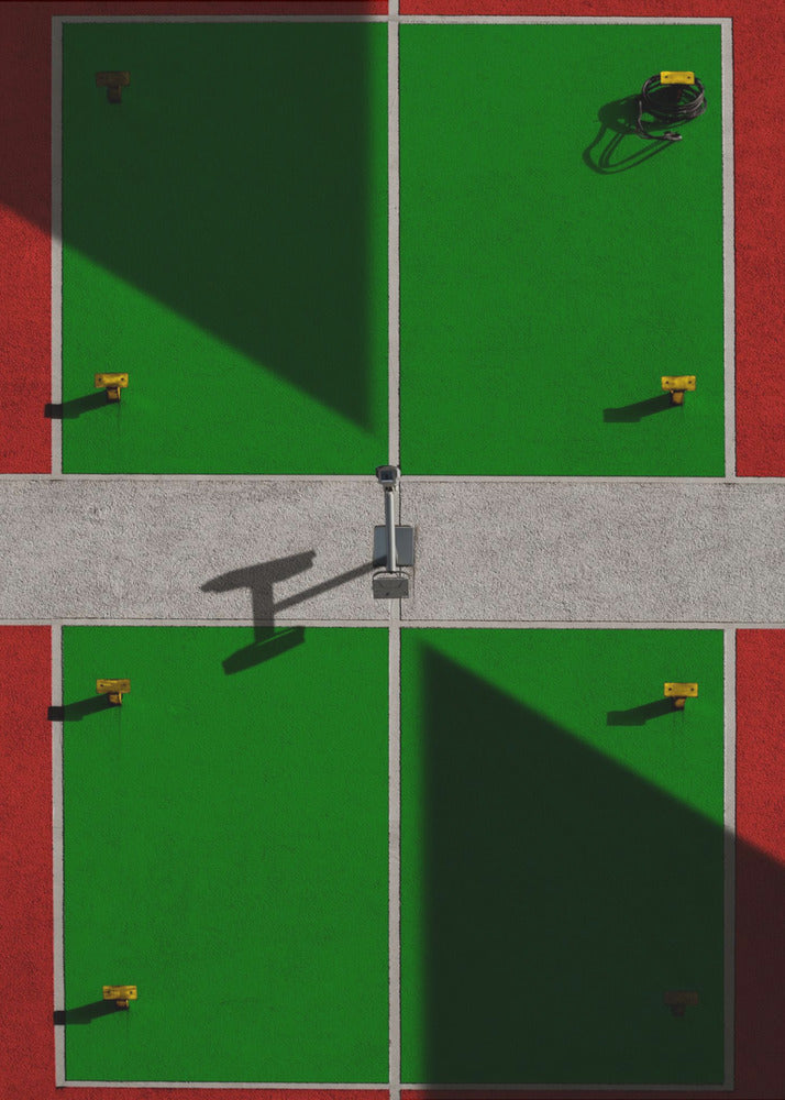 A high-angle, symmetrical photograph of a green and red sports court, viewed from directly above. Strong, sharp shadows cut diagonally across the composition, creating a geometric and abstract pattern of light and dark. A net post in the center casts a T-shaped shadow on the concrete dividing strip. Print