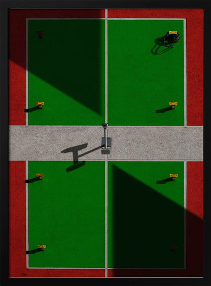 A high-angle, symmetrical photograph of a green and red sports court, viewed from directly above. Strong, sharp shadows cut diagonally across the composition, creating a geometric and abstract pattern of light and dark. A net post in the center casts a T-shaped shadow on the concrete dividing strip. Print