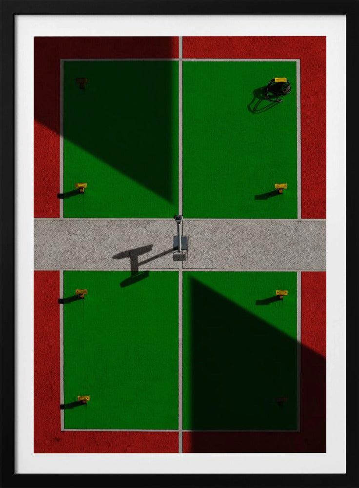 A high-angle, symmetrical photograph of a green and red sports court, viewed from directly above. Strong, sharp shadows cut diagonally across the composition, creating a geometric and abstract pattern of light and dark. A net post in the center casts a T-shaped shadow on the concrete dividing strip. Print