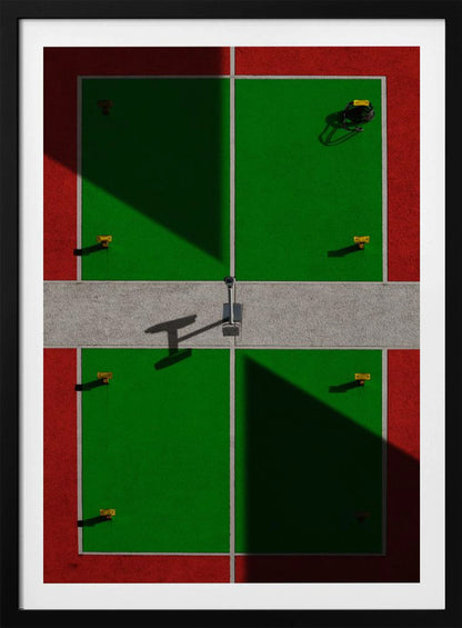 A high-angle, symmetrical photograph of a green and red sports court, viewed from directly above. Strong, sharp shadows cut diagonally across the composition, creating a geometric and abstract pattern of light and dark. A net post in the center casts a T-shaped shadow on the concrete dividing strip. Print