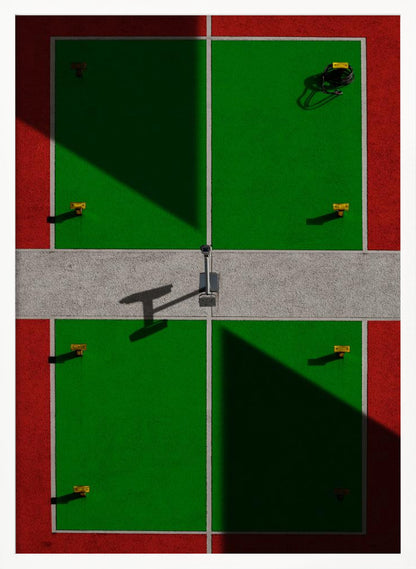 A high-angle, symmetrical photograph of a green and red sports court, viewed from directly above. Strong, sharp shadows cut diagonally across the composition, creating a geometric and abstract pattern of light and dark. A net post in the center casts a T-shaped shadow on the concrete dividing strip. Print