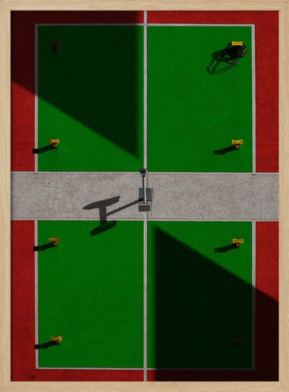 A high-angle, symmetrical photograph of a green and red sports court, viewed from directly above. Strong, sharp shadows cut diagonally across the composition, creating a geometric and abstract pattern of light and dark. A net post in the center casts a T-shaped shadow on the concrete dividing strip. Print