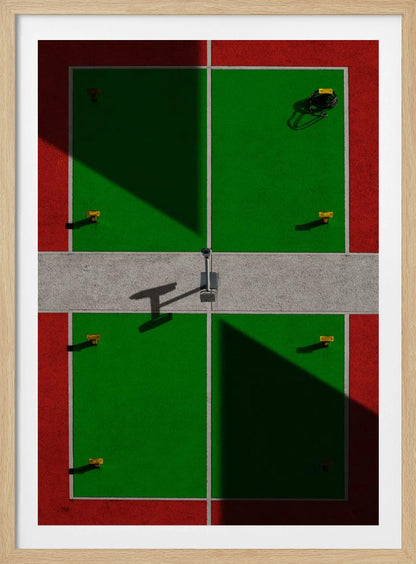 A high-angle, symmetrical photograph of a green and red sports court, viewed from directly above. Strong, sharp shadows cut diagonally across the composition, creating a geometric and abstract pattern of light and dark. A net post in the center casts a T-shaped shadow on the concrete dividing strip. Print