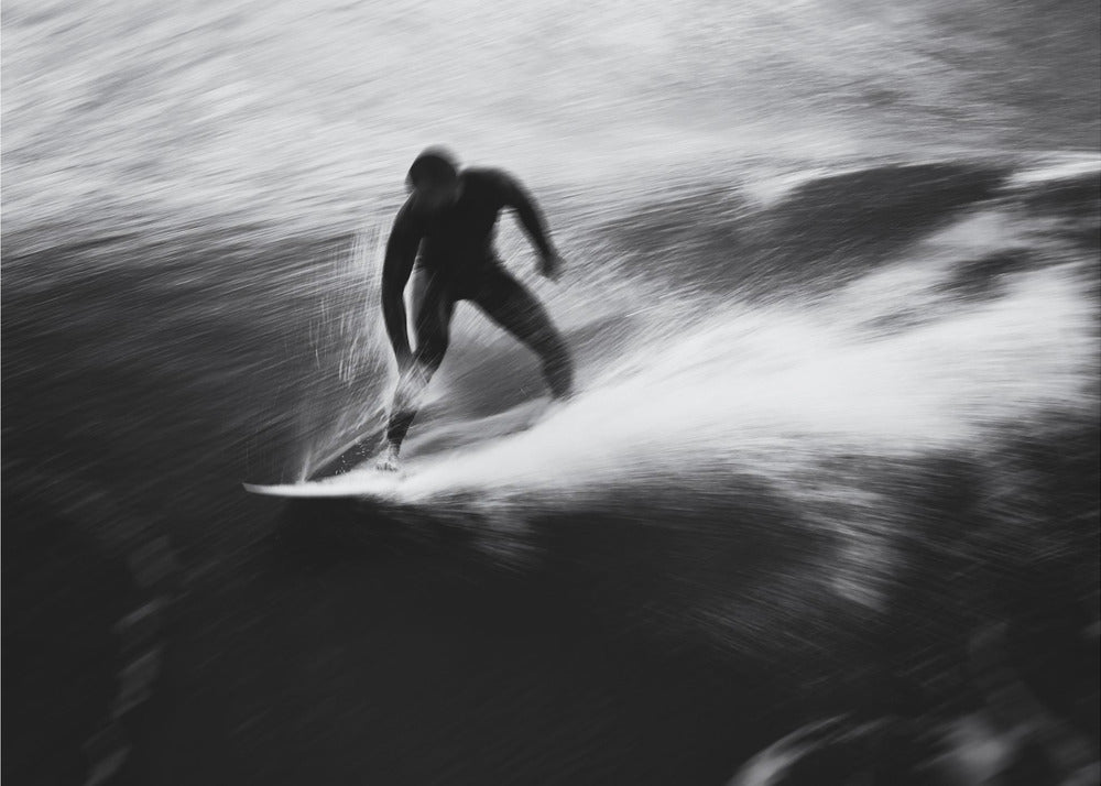 A dynamic black and white photograph of a surfer in a wetsuit carving through a wave. The image uses motion blur to emphasize the speed and energy of the water, creating a powerful and artistic effect. The artwork is presented in a simple silver frame. Print