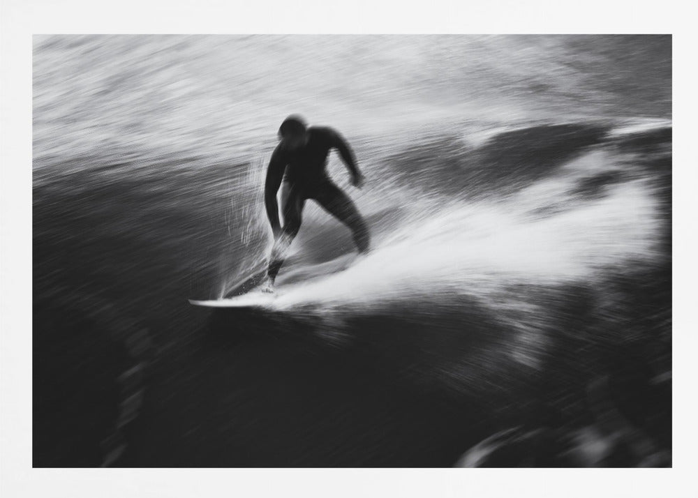A dynamic black and white photograph of a surfer in a wetsuit carving through a wave. The image uses motion blur to emphasize the speed and energy of the water, creating a powerful and artistic effect. The artwork is presented in a simple silver frame. Print