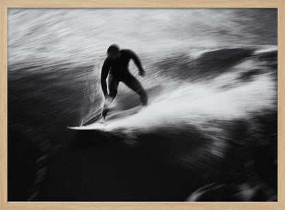 A dynamic black and white photograph of a surfer in a wetsuit carving through a wave. The image uses motion blur to emphasize the speed and energy of the water, creating a powerful and artistic effect. The artwork is presented in a simple silver frame. Print