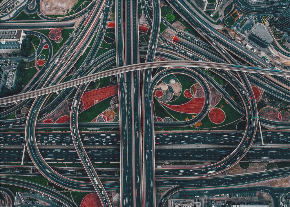 An aerial, top-down photograph of a sprawling and complex highway interchange, framed in silver. Multiple levels of roads and overpasses curve and intertwine, with blurred streaks of traffic visible. The spaces between the highways feature manicured green lawns and artistic red and patterned landscaping. Wall Art