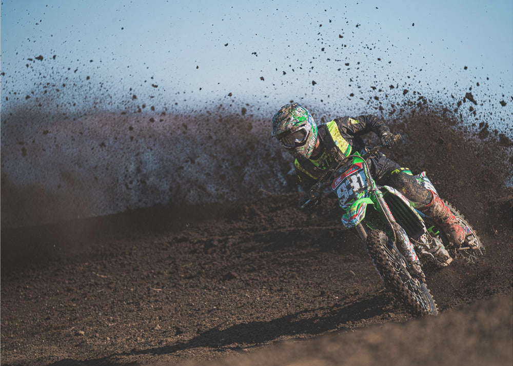 A motocross rider in green and black gear leans hard into a turn on a dirt track, kicking up a large spray of mud against a clear blue sky. Decor