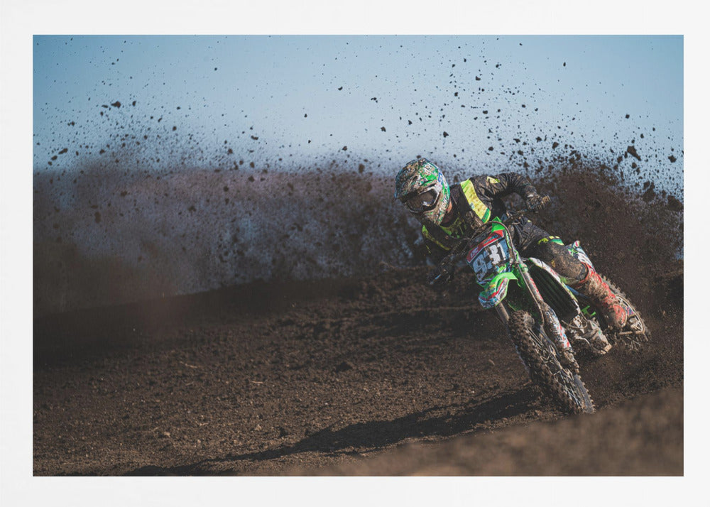 A motocross rider in green and black gear leans hard into a turn on a dirt track, kicking up a large spray of mud against a clear blue sky. Decor
