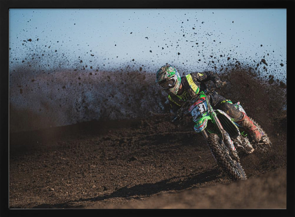 A motocross rider in green and black gear leans hard into a turn on a dirt track, kicking up a large spray of mud against a clear blue sky. Decor