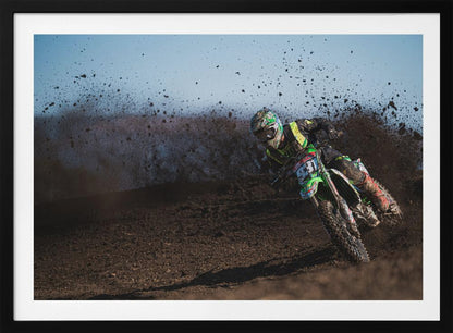 A motocross rider in green and black gear leans hard into a turn on a dirt track, kicking up a large spray of mud against a clear blue sky. Decor