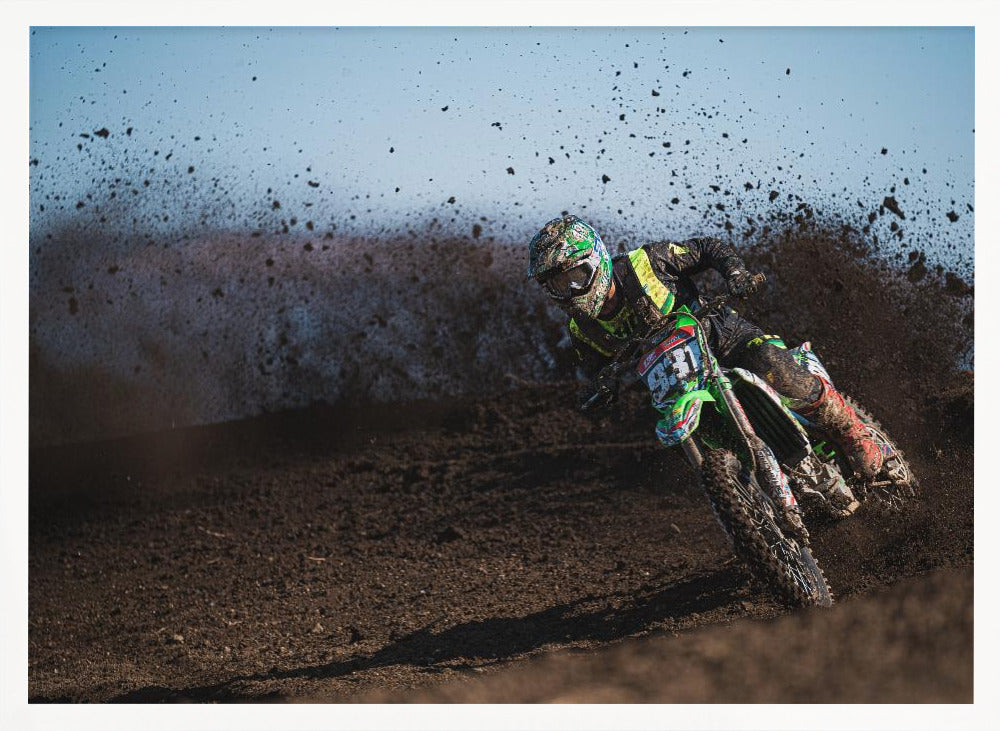 A motocross rider in green and black gear leans hard into a turn on a dirt track, kicking up a large spray of mud against a clear blue sky. Decor
