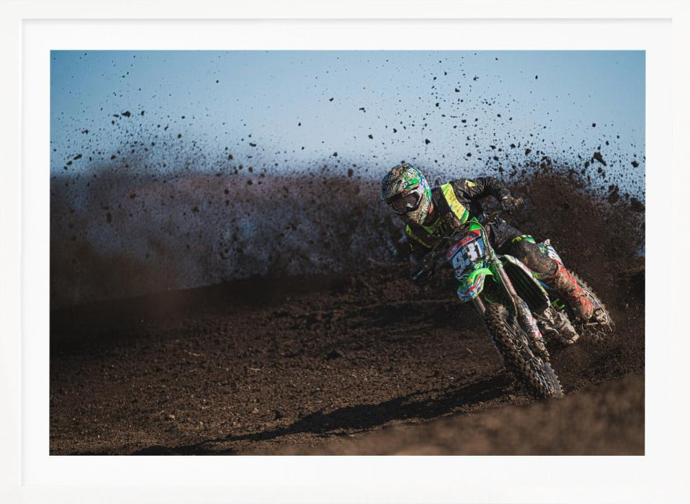 A motocross rider in green and black gear leans hard into a turn on a dirt track, kicking up a large spray of mud against a clear blue sky. Decor