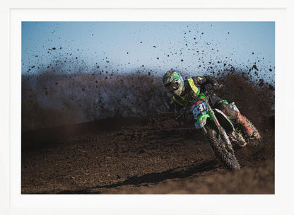 A motocross rider in green and black gear leans hard into a turn on a dirt track, kicking up a large spray of mud against a clear blue sky. Decor