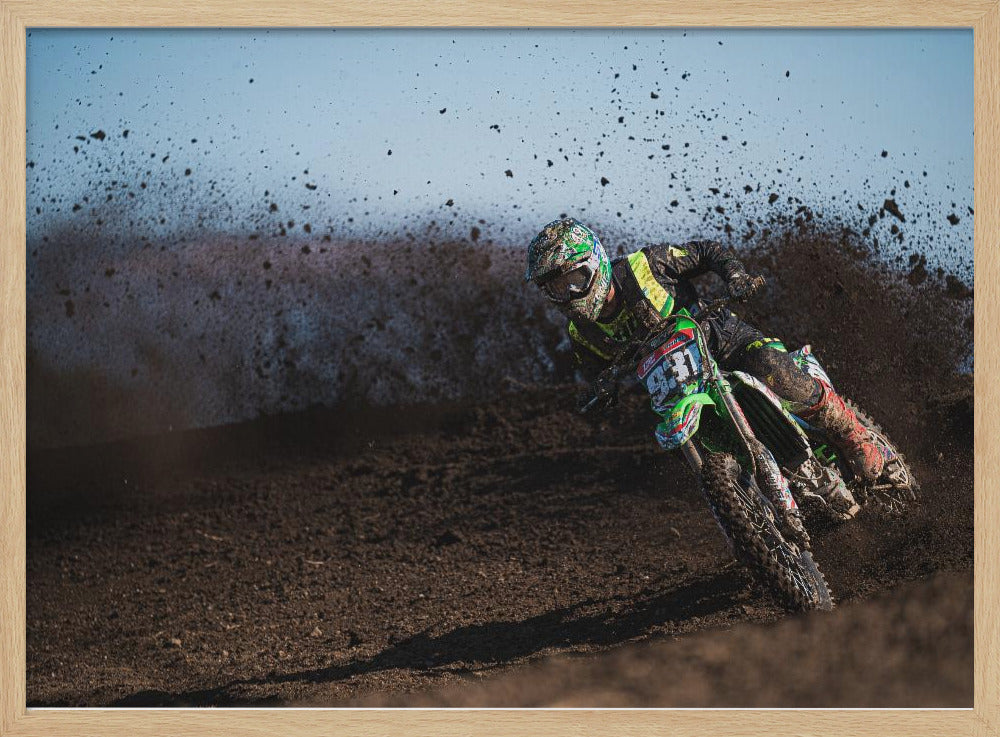 A motocross rider in green and black gear leans hard into a turn on a dirt track, kicking up a large spray of mud against a clear blue sky. Decor