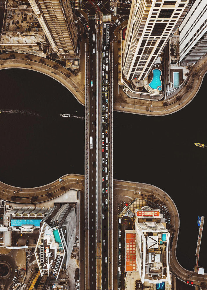 A symmetrical, top-down aerial photograph of a busy city bridge crossing dark water, flanked by towering skyscrapers with bright blue rooftop swimming pools. Print