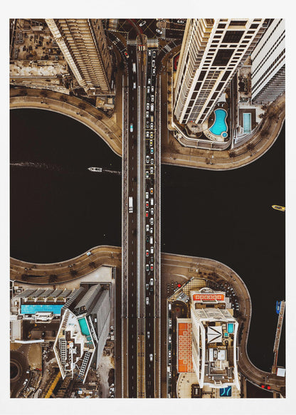 A symmetrical, top-down aerial photograph of a busy city bridge crossing dark water, flanked by towering skyscrapers with bright blue rooftop swimming pools. Print