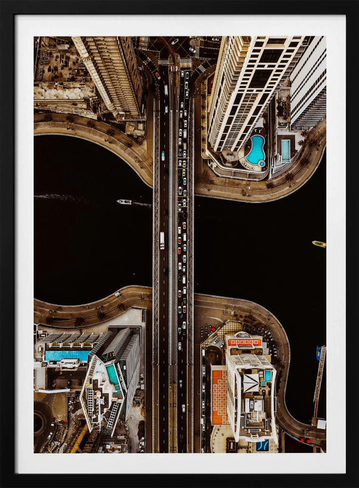 A symmetrical, top-down aerial photograph of a busy city bridge crossing dark water, flanked by towering skyscrapers with bright blue rooftop swimming pools. Print