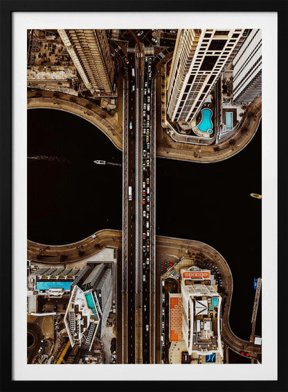 A symmetrical, top-down aerial photograph of a busy city bridge crossing dark water, flanked by towering skyscrapers with bright blue rooftop swimming pools. Print