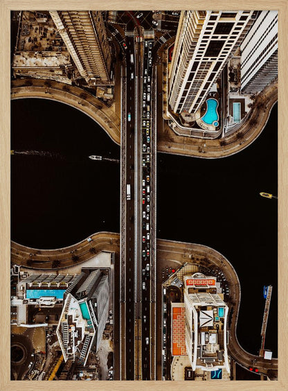 A symmetrical, top-down aerial photograph of a busy city bridge crossing dark water, flanked by towering skyscrapers with bright blue rooftop swimming pools. Print