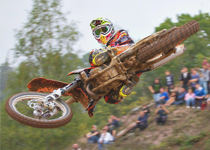 A dynamic action shot of a motocross racer in mid-air, leaning into a turn with their muddy orange dirt bike. The rider wears a colorful helmet and racing gear, with a blurred background of green trees and spectators watching from a dirt hill. The photo is enclosed in a silver frame. Decor