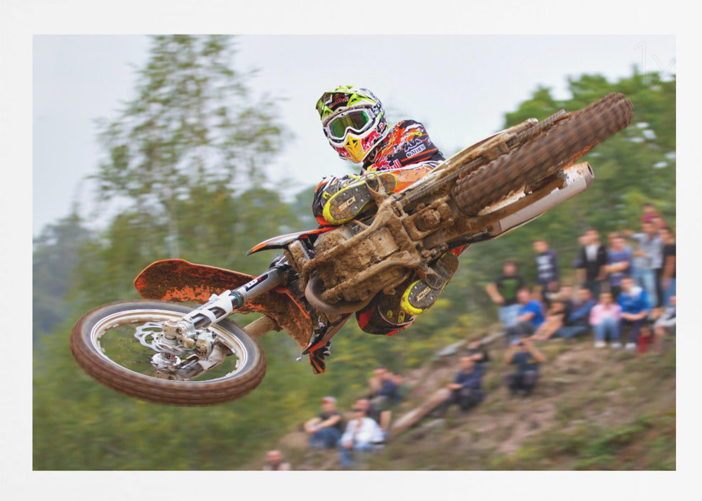 A dynamic action shot of a motocross racer in mid-air, leaning into a turn with their muddy orange dirt bike. The rider wears a colorful helmet and racing gear, with a blurred background of green trees and spectators watching from a dirt hill. The photo is enclosed in a silver frame. Decor