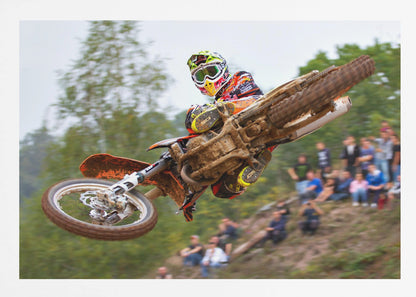 A dynamic action shot of a motocross racer in mid-air, leaning into a turn with their muddy orange dirt bike. The rider wears a colorful helmet and racing gear, with a blurred background of green trees and spectators watching from a dirt hill. The photo is enclosed in a silver frame. Decor