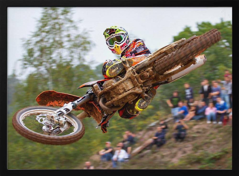 A dynamic action shot of a motocross racer in mid-air, leaning into a turn with their muddy orange dirt bike. The rider wears a colorful helmet and racing gear, with a blurred background of green trees and spectators watching from a dirt hill. The photo is enclosed in a silver frame. Decor